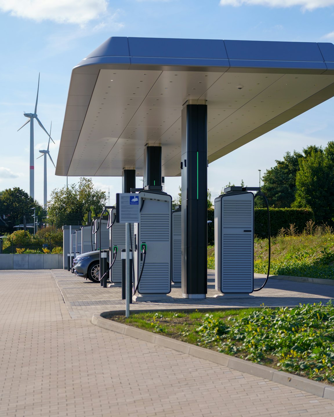 Elektriautode laadijad varikatuse all, tuuleturbiinid tagaplaanil.